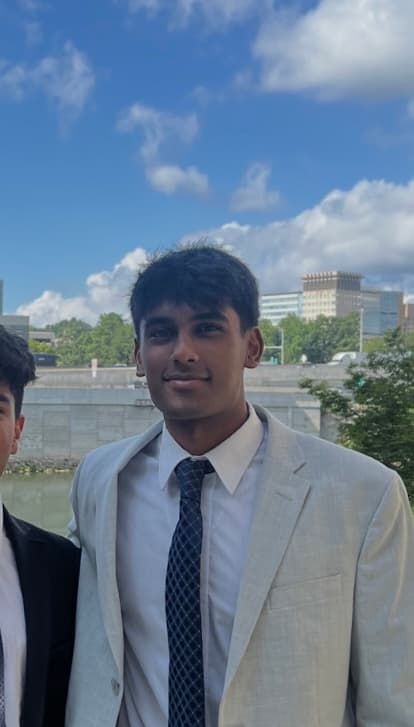 Smiling young man in a light grey suit and blue tie with city buildings behind.